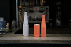On a coffee counter, two Duet products are shown side by side. One grey Duet is fully assembled with the coffee cup stacked on the water bottle. The second coral Duet shows the coffee cup and water bottle with their lids attached, positioned next to each other.